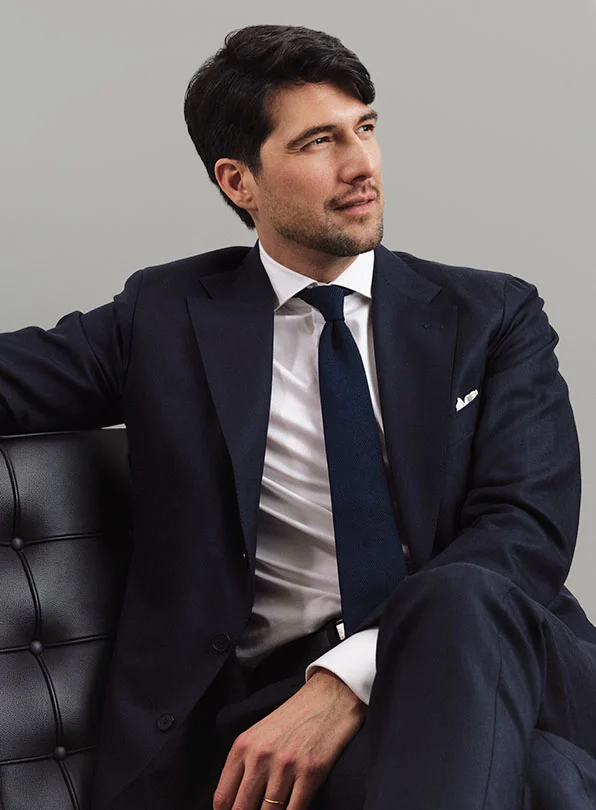 Man with dark hair in a black suit and tie, sitting on the black leather chair with legs crossed, looking to the side against a light gray background - Male procedures by Dr. Emily Deane