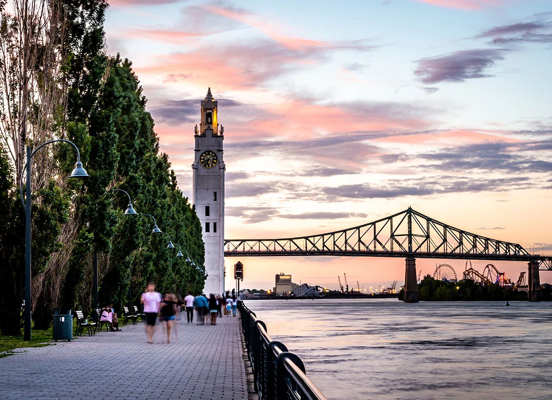 Peaceful waterfront promenade lined with trees featuring a historic clock tower and bridge during a colorful sunset. - Out of town in Montreal, QC