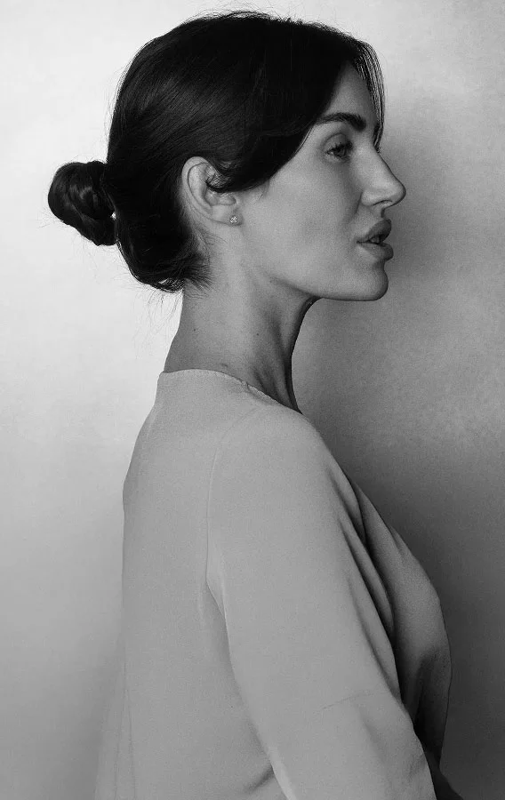 Woman in profile with hair in low bun wearing neutral toned shirt against white wall - in Montreal, QC